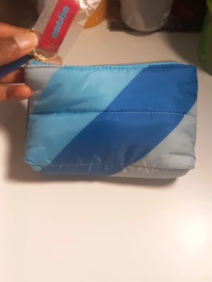 Blå vadderad necessär Estée Lauder - Säljer en vadderad necessär från Estée Lauder i olika blå nyanser med diagonal design. Väskan har dragkedja upptill och är tillverkad i ett glansigt syntetmaterial. Perfekt för smink eller småprylar när du är på språng.