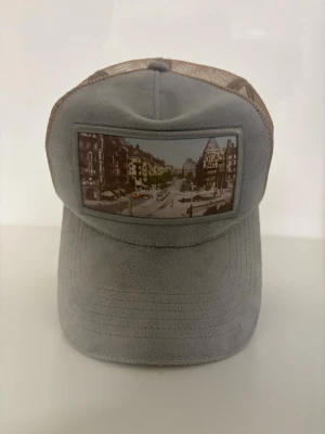 Grå Garavelli keps  - Snygg grå keps från Garavelli med ett coolt stadsmotiv framtill. Kepsen har mesh-bakstycke i beige och böjd skärm. Perfekt accessoar för en urban look och passar till många outfits.