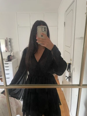 Svart omlottklänning med volang - Säljer en svart kort omlottklänning med v-ringning och långa, vida ärmar. Klänningen har volangdetaljer i kjolen och ger en flowy känsla. Perfekt för dig som gillar en enkel men snygg look. Den är helt oanvänd med lapp kvar