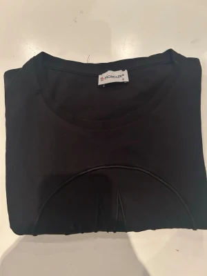 Svart Moncler t-shirt med logga - Snygg svart t-shirt från Moncler med stor ton-i-ton logga på bröstet. Klassisk rund halsringning och korta ärmar. Tillverkad i mjuk bomull som känns skön mot huden. Perfekt för dig som gillar stilrena plagg med exklusiv känsla.