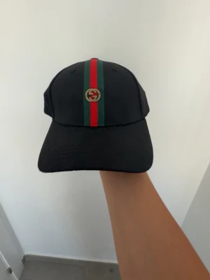 Svart keps med grön röd detalj  - Snygg svart keps  med klassisk röd och grön rand längs mitten  Kepsen har böjd skärm.perfekt accessoar för dig som gillar streetwear och lyx.
