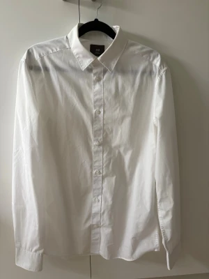 Vit slim fit skjorta från H&M - Klassisk vit skjorta från H&M i slim fit-modell. Skjortan har långa ärmar, knappar framtill och en stilren krage. Tillverkad i ett skönt material som är lätt att stryka
