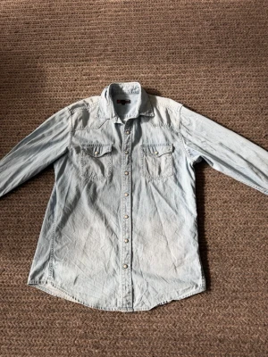 Ljusblå jeansskjorta med tryckknappar - Säljer en ljusblå jeansskjorta i slim fit med klassisk krage och två bröstfickor med lock. Skjortan har tryckknappar framtill och på fickorna, samt långa ärmar. Perfekt för en avslappnad och cool stil.