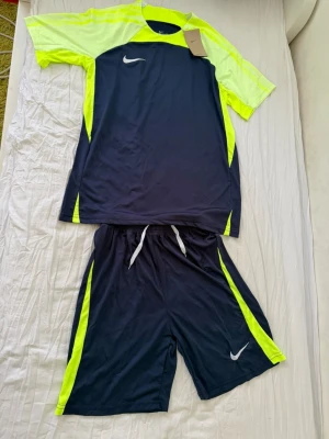 Nike sportset marinblå/gul - Snyggt sportset från Nike med t-shirt och shorts i marinblått och neongult. Tillverkad i lätt och snabbtorkande material, perfekt för träning eller match. T-shirten har rund hals och korta ärmar, shortsen har dragsko i midjan för bästa passform.