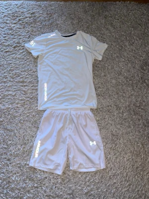Vita träningsset Under Armour - Vit träningsset från Under Armour med elastisk midja och logga på vänster ben. Shortsen och tröjan är gjorda i ett lätt och snabbtorkande syntetmaterial, perfekta för sport och aktiva dagar. Loggorna och text lyser även i blixt!