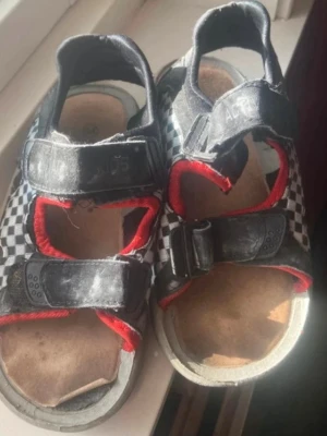 Schackrutiga sandaler med kardborre(barn)  - Svarta sandaler med vitt schackrutigt mönster och röda detaljer. De har två kardborreband och en öppen tådesign. Sulan är beige och insidan är vadderad för extra komfort. Perfekt för sommardagar och enkel att ta på.