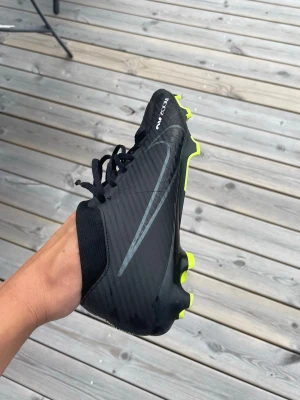 Nike svarta fotbollsskor med neon - Säljer ett par svarta Nike air zoom fotbollsskor med snygga neonfärgade dobbar och detaljer. Skorna har en modern design med snörning och en strumpa som går upp över ankeln. Materialet är syntetiskt och väldigt skönt.