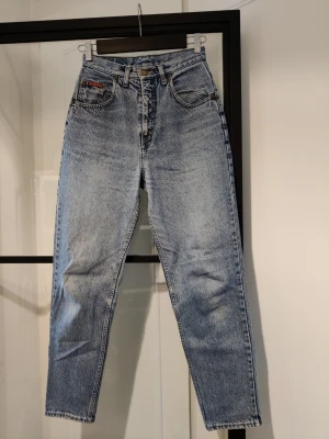 Blå mom jeans med hög midja - Ett par klassiska blå mom jeans med hög midja och raka ben. Jeansen har fem fickor, knappgylf och en vintage tvättad look. Materialet är kraftig denim i 100% bomull. Perfekta för en avslappnad och trendig stil.