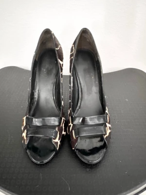Bruna pumps med giraffmönster - Snygga pumps med giraffmönstrad ovandel i brunt och beige. Skorna har öppen tå och svarta lackdetaljer framtill. Klackarna är höga och smala, vilket ger en elegant siluett. Perfekta för dig som vill sticka ut med ett djurmönstrat statement.
