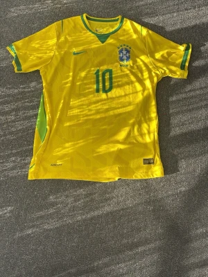 Brasilien Neymar 10 matchtröja - Gul Brasilien fotbollströja från Nike med gröna detaljer, Neymar Jr och nummer 10 på ryggen. Tröjan har rund hals, korta ärmar och är tillverkad i lätt och ventilerande material med Aerofit-teknologi. Perfekt för dig som älskar fotboll och vill bära landslagets färger.
