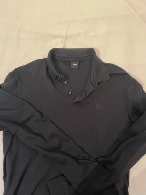 Svart långärmad piké från Hugo Boss - Klassisk svart långärmad pikétröja från Hugo Boss med diskret broderad logga på bröstet. Tröjan har krage, knäppning med tre knappar och är tillverkad i mjukt material som ger en clean och stilren look.