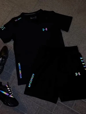 Svart träningsset från Under Armour - Snyggt svart träningsset från Under Armour med t-shirt och shorts. Loggor och text i regnbågsfärger ger en modern vibe. Perfekt för gymmet eller löprundan. Materialet är lätt och andas, vilket gör det bekvämt under träning.