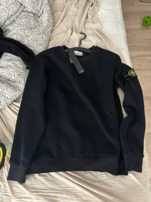 Svart sweatshirt från Stone Island - Svart sweatshirt från Stone Island med klassisk rund hals och ribbade muddar. Ikonisk logopatch på vänster ärm. Perfekt för dig som gillar streetwear och enkel stil.