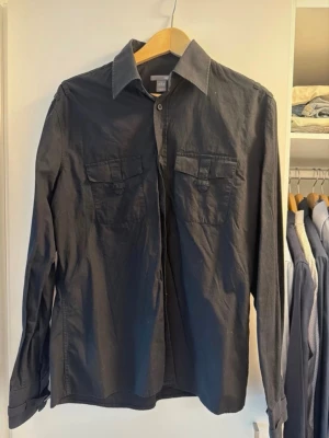 Svart skjorta med bröstfickor medium - Stilren svart skjorta i bomull med klassisk krage och två bröstfickor med lock. Skjortan har långa ärmar och knappar framtill. Perfekt för en clean och enkel look. Medium