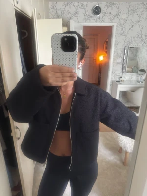 Svart teddyjacka med dragkedja - Mysig svart teddyjacka med krage och hel dragkedja framtill. Jackan har en boxy passform och två stora fickor framtill. Perfekt för att hålla värmen och ge en trendig look till din outfit.
