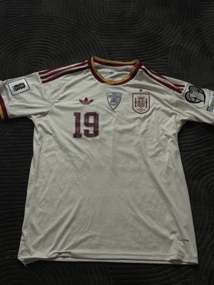 Vit Spanien fotbollströja nr 19 - Säljer en vit Spanien fotbollströja från Adidas med vinröda detaljer och nummer 19. Tröjan har officiella märken, klubbmärke och trycket 'LAMINE YAMAL' på ryggen. Tillverkad i lätt och ventilerande material, perfekt för match eller träning.