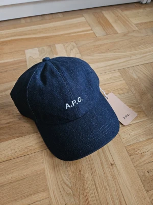 Mörkblå denimkeps från A.P.C. - Snygg mörkblå keps i denim från A.P.C. med vit broderad logga framtill. Klassisk böjd skärm och justerbar rem bak. Perfekt för dig som gillar minimalistisk och stilren streetwear.