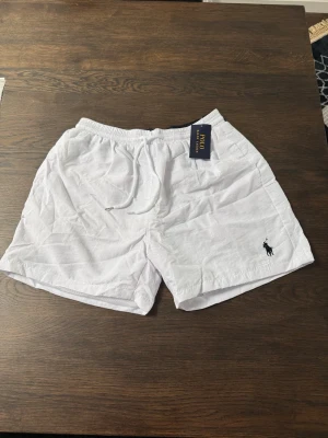 Vita shorts från Polo Ralph Lauren - Snygga vita shorts från Polo Ralph Lauren med klassisk broderad logga i svart på benet. Shortsen har elastisk midja med snörning och är tillverkade i ett lätt bomullstyg, perfekta för varma dagar.