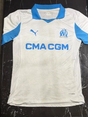 Marseille matchtröja Puma vit/blå - Snygg Marseille matchtröja från Puma i vitt med blå detaljer. Tröjan har klubbmärke och Puma-logga på bröstet samt sponsortryck 'CMA CGM' framtill. Tillverkad i lätt och ventilerande material, perfekt för träning eller match.