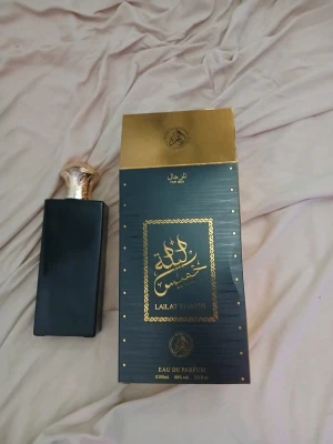 Lailat Khamees Eau de Parfum 100ml - Elegant parfym med svart, rektangulär flaska och dekorativ guldfärgad kork. Kommer i lyxig svart och guld förpackning med arabiska detaljer. Doften är Eau de Parfum och flaskan rymmer 100 ml.