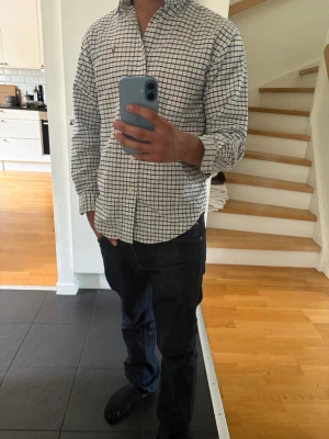 Rutig skjorta från Polo Ralph Lauren - Klassisk vit skjorta med mörkblå rutmönster från Polo Ralph Lauren. Skjortan har lång ärm, knappar framtill och den ikoniska broderade loggan på bröstet. Perfekt till jeans eller chinos för en clean och stilren look.