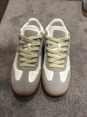 Vita och grå sneakers med mockadetaljer - Snygga sneakers i vitt och ljusgrått med beige skosnören och detaljer i mocka. Yttermaterialet är en mix av slätt läder och mocka, och sulan är i klassisk gummifärg. Perfekta för en clean och stilren look. Brandless shoes. 