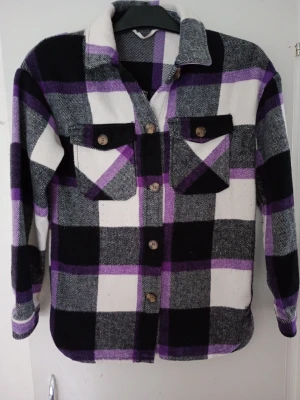 Rutig overshirt i svart, vit och lila - Snygg overshirt med rutigt mönster i svart, vit, grå och lila. Två bröstfickor med lock och knappar, klassisk krage och knäppning framtill. Tillverkad i ett mjukt, tjockare tyg som ger en avslappnad look. Perfekt lager på lager-plagg.