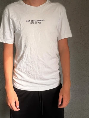 Vit t-shirt med texttryck - Vit t-shirt med svart texttryck 'LOW EXPECTATIONS HIGH HOPES' på bröstet. Klassisk passform med rund hals och korta ärmar. Tillverkad i mjuk bomull som känns skön mot huden. Perfekt till jeans eller shorts för en enkel och clean look.
