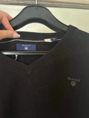 Svart stickad tröja från GANT  - Klassisk svart stickad tröja från GANT med v-ringning och diskret logga broderad på bröstet. Tröjan har långa ärmar och ribbade muddar vid ärmslut och nederkant. Perfekt till alla tillfällen och enkel att matcha.