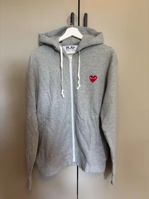 CDG Play Hoodie - Säljer en grå zip-hoodie med huva och vita snören. På bröstet sitter ett litet rött hjärta med ögon, vilket ger en unik och lekfull touch. Hoodien har långa ärmar och är tillverkad i mjukt material, perfekt för en chill och avslappnad stil.