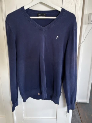 Mörkblå V-ringad PeakPerformance  - En stilren mörkblå V-ringad tröja med lång ärm och ribbade muddar. Tröjan har en liten vit broderad logga på bröstet och är tillverkad i ett mjukt material som passar perfekt till jeans eller chinos. Skriv om ni har några frågor! 