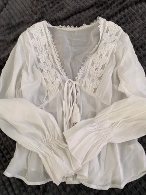 Vit boho-blus med spetsdetaljer - Säljer en romantisk vit blus i tunn, luftig bomull med v-ringning och breda spetsdetaljer längs halsen. Blusen har långa ärmar med smock vid handlederna och knytband framtill. Perfekt för en bohemisk look och superfin till jeans eller kjol.