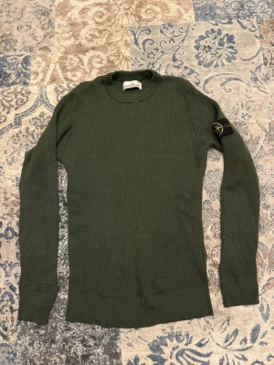 Grön stickad tröja från Stone Island - Mörkgrön ribbstickad tröja från Stone Island med klassisk patch på vänster ärm. Tröjan har rund halsringning, långa ärmar och breda muddar vid ärmslut och nederkant. Perfekt för dig som gillar streetwear och snygga detaljer.