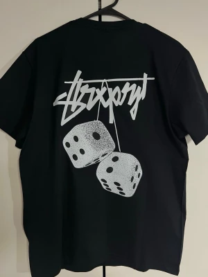 Svart Stüssy t-shirt med tärningsmotiv - Svart t-shirt från Stüssy med stort tryck av två tärningar och grafisk text på ryggen samt mindre logga och text på bröstet. Klassisk passform och rund halsringning. Perfekt för dig som gillar streetwear och snygga prints.