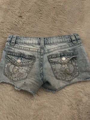 Blå jeansshorts med broderade fickor - Snygga blå jeansshorts med slitna kanter och broderade detaljer på bakfickorna. Shortsen har knappstängning på fickorna och en klassisk femficksmodell. Perfekta för varma dagar och ger en avslappnad look.