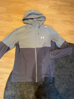 Grå vindjacka från Under Armour - Underarmour jacka, ihop sydda hål på ena armen syns knappt när man har på sig den. Väldigt bra skick inte använd så mycket. Pris kan diskuteras, skirv om du har några frågor!