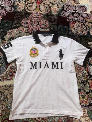 Vit piké från Polo Ralph Lauren - Vit pikétröja från Polo Ralph Lauren med svart krage och detaljer. Stor broderad logga på bröstet, MIAMI-tryck och emblem. Svarta detaljer på ärmarna och nummer 3 på ena ärmen. Klassisk och sportig stil.