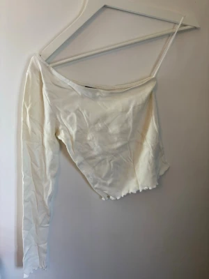 Vit oneshoulder topp med lång ärm - Säljer en vit oneshoulder topp med en lång ärm. Tillverkad i ribbad bomull som ger en snygg och bekväm passform. Väldigt fint skick från gina tricot.