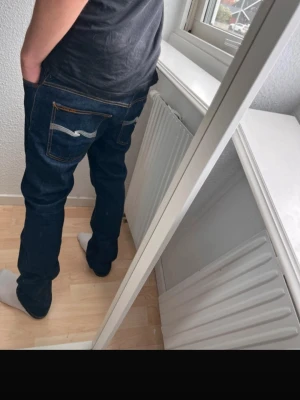 Mörkblå jeans från Nudie Jeans - Säljer ett par mörkblå jeans från Nudie Jeans med klassisk femficksmodell och kontrastsömmar. Modellen är Thin Finn. Jeansen har en rak passform och den ikoniska Nudie-bågen på bakfickorna. Tillverkade i slitstarkt denimtyg, perfekta för en avslappnad stil.