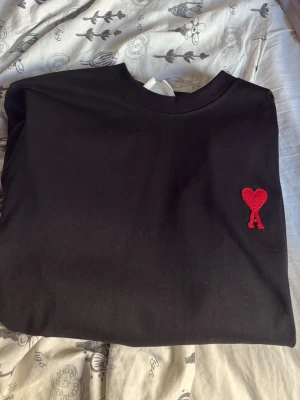 Svart t-shirt från Ami Paris - Säljer en svart t-shirt från Ami Paris med det ikoniska röda hjärtat och A-broderiet på bröstet. Tröjan har rund halsringning och är gjord i mjukt bomullsmaterial. Enkel och stilren design som passar till allt.