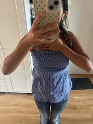 Blåvit randig ärmlös topp - Säljer en söt blåvit randig topp med knappar framtill och smala axelband. Toppen har en lätt utsvängd passform och är perfekt för varma dagar. Stilen är enkel men trendig och passar till jeans eller kjol.