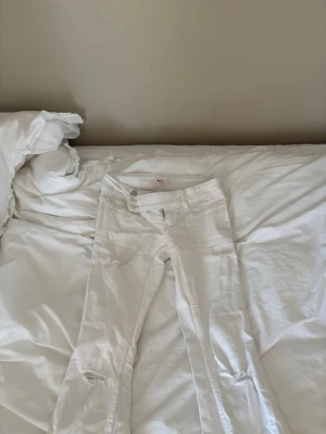 Vita bootcut jeans med broderi - Säljer ett par vita bootcut jeans med snygga slitningar vid knäna och detaljerat broderi på bakfickorna. Jeansen har låg midja och dubbla knappar framtill. Perfekta för dig som vill sticka ut lite extra! ÄLSKAR dessa jeans men dem kommer dock aldrig till användning tyvärr🥰