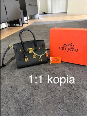 Svart Birkin handväska med gulddetaljer - Stilren svart handväska i strukturerat material med guldfärgade detaljer, kedja och hänglås. Väskan har både handtag och axelrem för olika bärstilar. Klassisk fyrkantig form och dekorativa berlocker framtill.