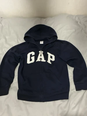Mörkblå hoodie med dragkedja från GAP - Mörkblå hoodie från GAP med vit broderad logga på bröstet. Tröjan har dragkedja framtill, huva och ribbade muddar vid ärmslut och nederkant. Tillverkad med minst 20% återvunnet material. Perfekt för en avslappnad och stilren look.