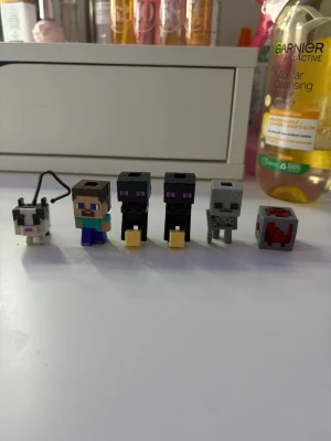 Kinder Minecraft minifigurer och block - Säljer ett set med sex små Minecraft-figurer i plast. Innehåller Steve, två Endermans, en katt, ett skelett och ett rött block. Figurerna är fyrkantiga och har klassiska pixelgrafikfärger i svart, grått, blått, grönt och rött. Perfekt för samlare eller som dekoration. 