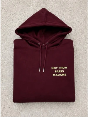 Vinröd not from Paris madame hoodie herr - Säljer en vinröd hoodie med huva och dragsko. På bröstet och ryggen står det 'NOT FROM PARIS MADAME' i guld text. Hoodien har en klassisk passform och är gjord i mjukt material, perfekt för en chill och trendig look.