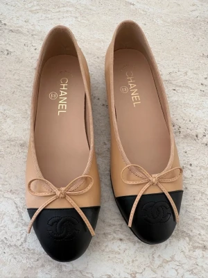Beige och svarta ballerinaskor Chanel - Snygga ballerinaskor från Chanel i beige läder med svart tåparti och klassisk rosett framtill. Skorna har rundad tå och den ikoniska Chanel-loggan präglad på tån. Perfekt för dig som gillar tidlös och elegant stil. Storlek 37. Låda ingår ej.
