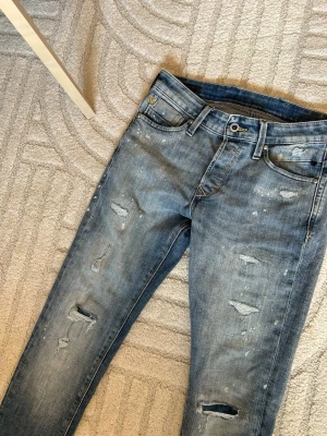 Blå slitna jeans 28/32 - Säljer ett par blå skinny jeans med slitningar och distressed detaljer på både fram- och baksida. Jeansen har klassisk femficksmodell, bälteshällor och dragkedjegylf. Perfekta för en avslappnad och trendig look.