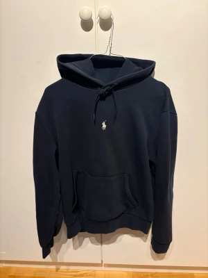 Mörkblå Ralph Lauren hoodie  - Snygg mörkblå hoodie med huva och dragsko samt en stor magficka framtill. Hoodien har ribbade muddar vid ärmslut och nederkant. Perfekt för en avslappnad och stilren look.