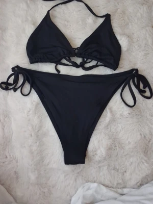 Svart bikini med knyt - Snygg svart bikini med trekantsöverdel och knyt både i nacken och ryggen. Underdelen har höga ben och knyt i sidorna för justerbar passform. Perfekt för strandhäng eller poolparty.